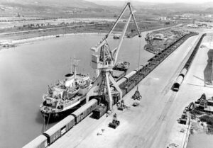 Foto d'epoca in bianco e nero delle prime banchine operative del porto di Capodistria (Luka Koper) con gru a portale e treni merci.